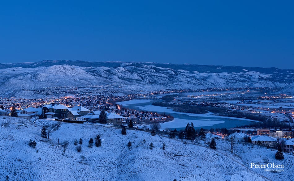 blue sky with snowy hills and ground with trees and water with lit city