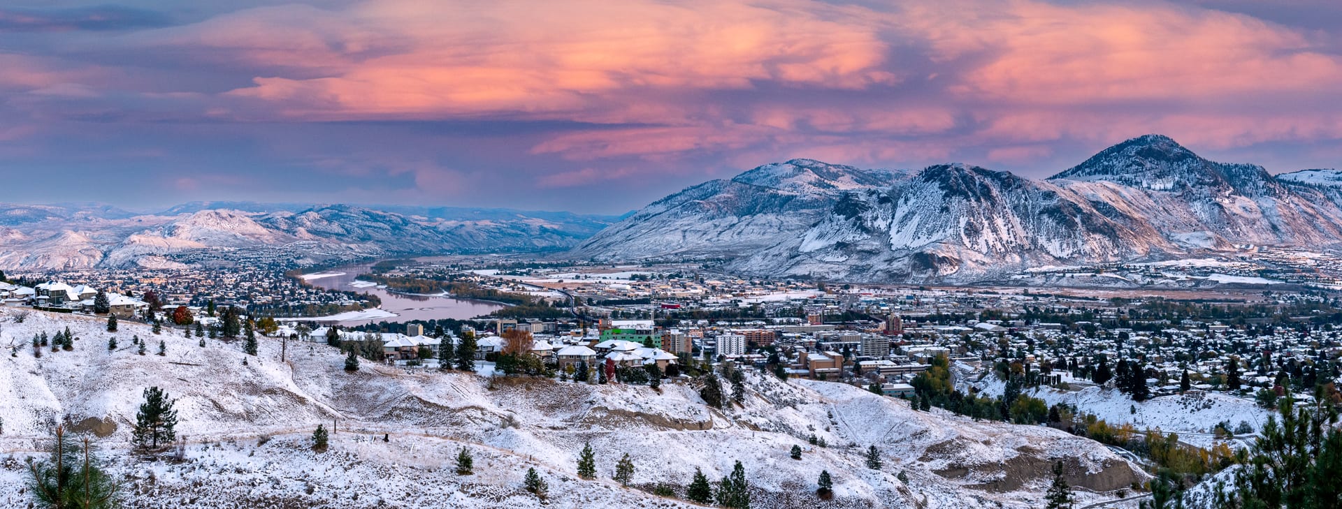 snowy mountain view with city and trees with pink and blue clouds