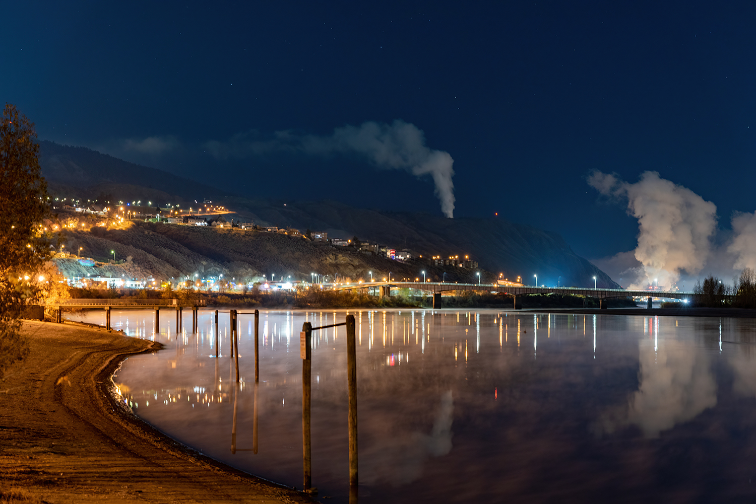 lakeside view of a city at night with smoke and mountains